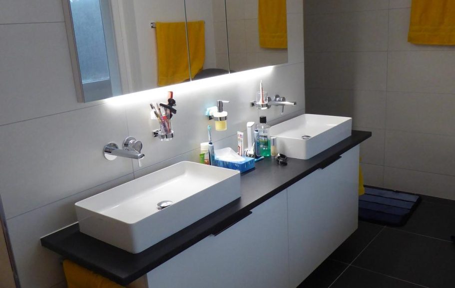 Zwei weiße Waschbecken auf einem schwarzen Tisch, mit Hygieneartikeln und gelben Handtüchern.